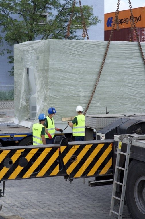 Rozładunek prefabrykatu stacji transformatorowej dźwigiem — element podwieszony nad placem budowy